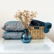 Itens decorativos de casa sobre móvel branco, incluindo duas almofadas azuis, manta azul enrolada em cesto de palha e vaso de vidro azul com arranjo de flores secas.