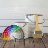 Latas de tinta metálicas de diferentes tamanhos dispostas sobre piso de madeira, com leque de amostras de cores e pincel de cabo de madeira apoiado ao lado.