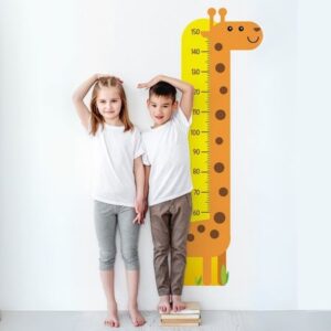 uma menina loira marcando na parede a sua altura, ao lado de um menino menor que ela, que está em cima de alguns livros para conseguir a mesma altura dela. Na parede, atrás deles, tem uma girafa que marca a altura da criança, para acompanhar seu desenvolvimento
