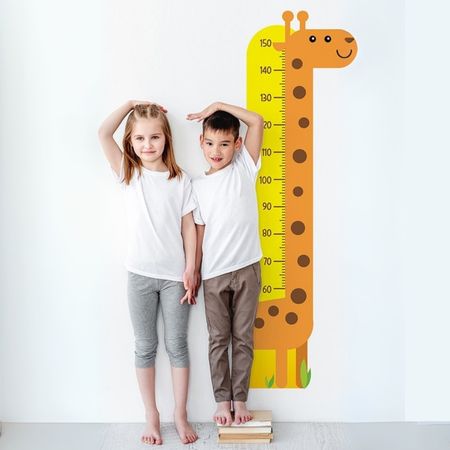 uma menina loira marcando na parede a sua altura, ao lado de um menino menor que ela, que está em cima de alguns livros para conseguir a mesma altura dela. Na parede, atrás deles, tem uma girafa que marca a altura da criança, para acompanhar seu desenvolvimento
